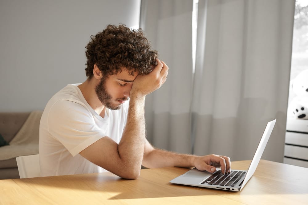 Young man feeling fatigued with too many surveys on a laptop with a hand on his head.
